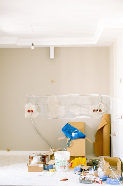 A close-up view of a silver metal folding hand truck with three horizontal ridged platforms used for transporting boxes, positioned indoors against a plain white wall. Behind the hand truck, there are