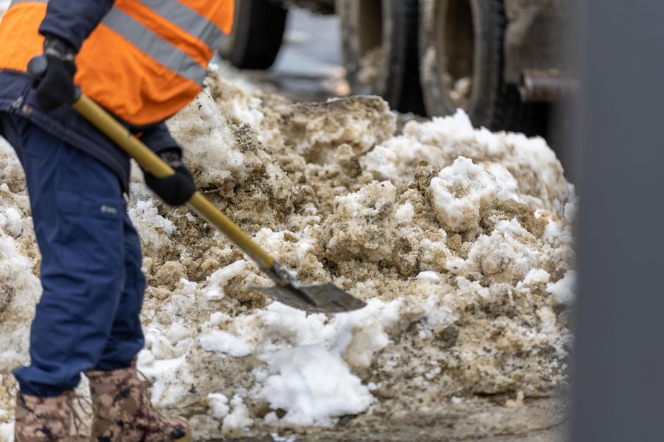 A person wearing an orange high-visibility vest, blue work trousers, and patterned boots is shovelling a mixture of snow, ice, and dirt on a pavement outside a property. The individual is using a yell
