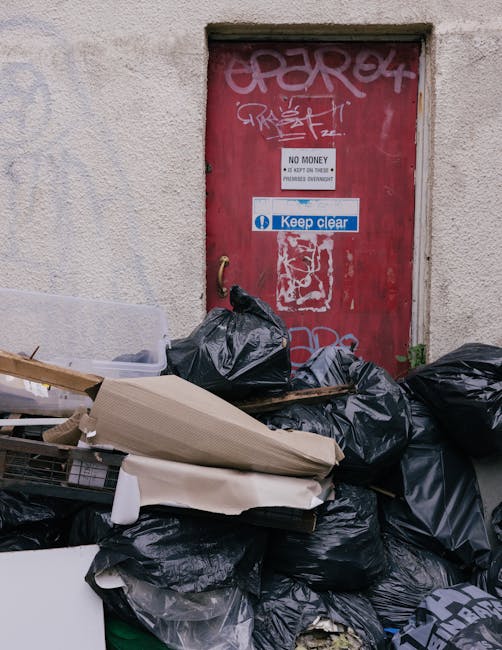 The image depicts a narrow alleyway closed off by a weathered, red metal door with graffiti and multiple signs, including a white 'No Money' notice and a blue 'Keep Clear' sign. In front of the door, 