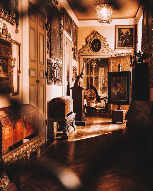 A spacious, ornately decorated room with antique furniture and elaborate wall moldings, illuminated by warm lighting from a chandelier and possible natural light. The walls are adorned with numerous f