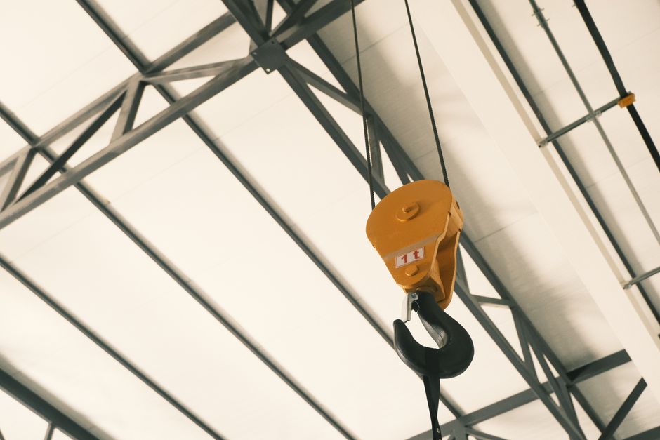 A close-up view of a yellow specialist lifting hoist with a maximum capacity of 1 tonne, suspended from a metal scaffolding structure inside a building. The hoist has a large, curved black hook hangin